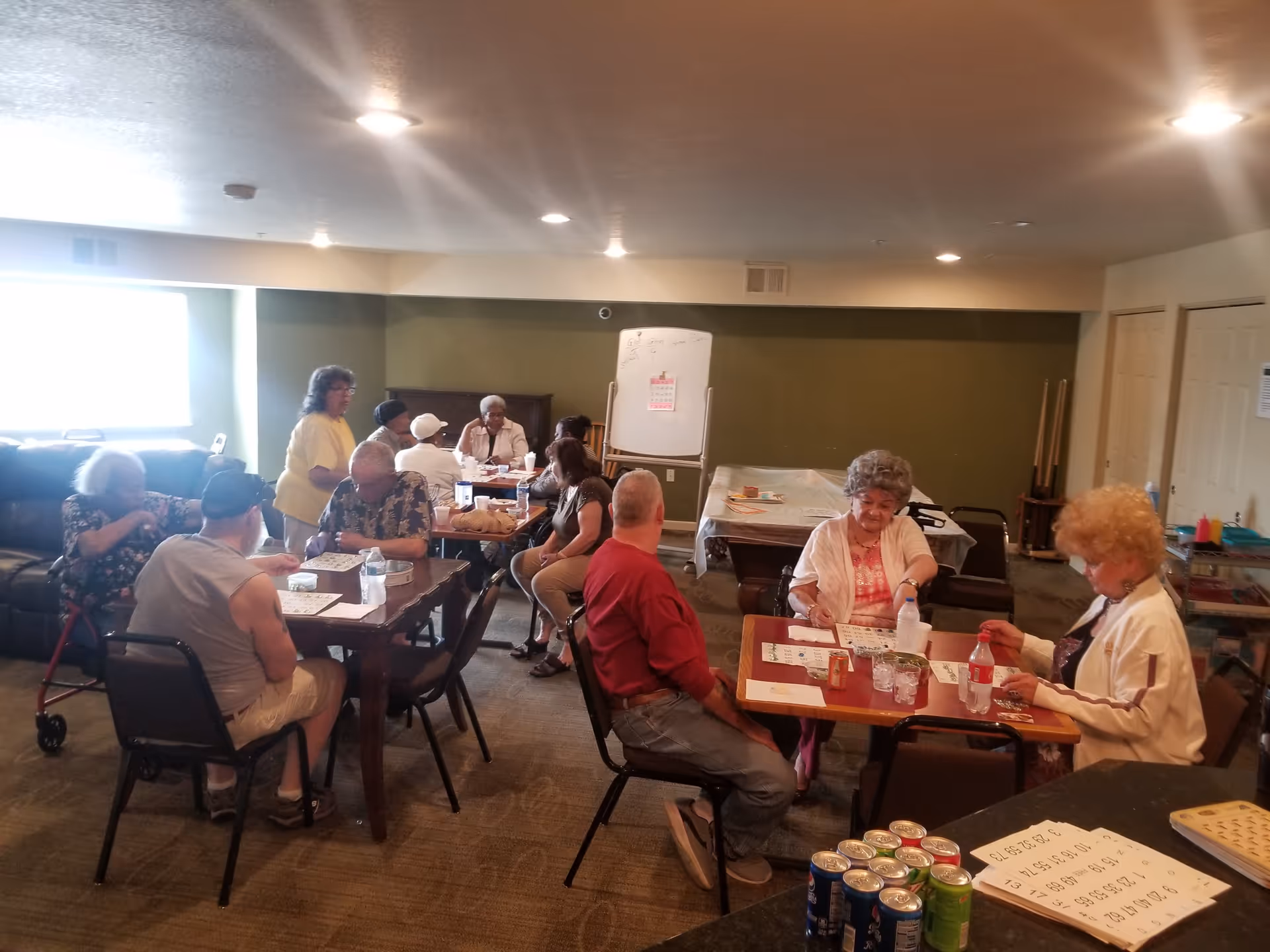 A group of elderly people sitting around tables in a common room playing bingo. There are bingo cards, drinks, and snacks on the tables. The room has a green accent wall, a whiteboard, and a pool table covered with a cloth in the background. The atmosphere appears social and relaxed.