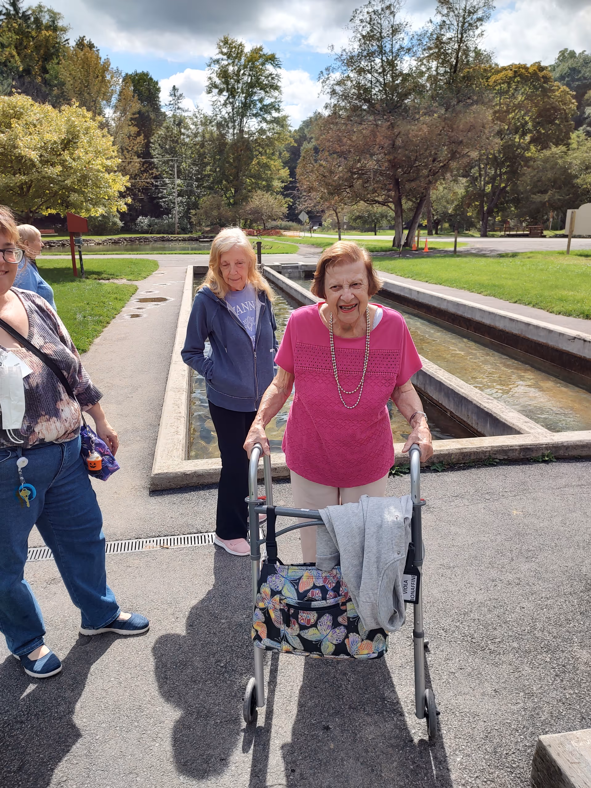 Three elderly women outdoors near a narrow water feature with trees and grass in the background. One woman in a pink shirt is using a walker with a butterfly-patterned bag attached, smiling at the camera. Another woman in a blue jacket stands behind her, and a third woman in a patterned top and jeans stands to the left.