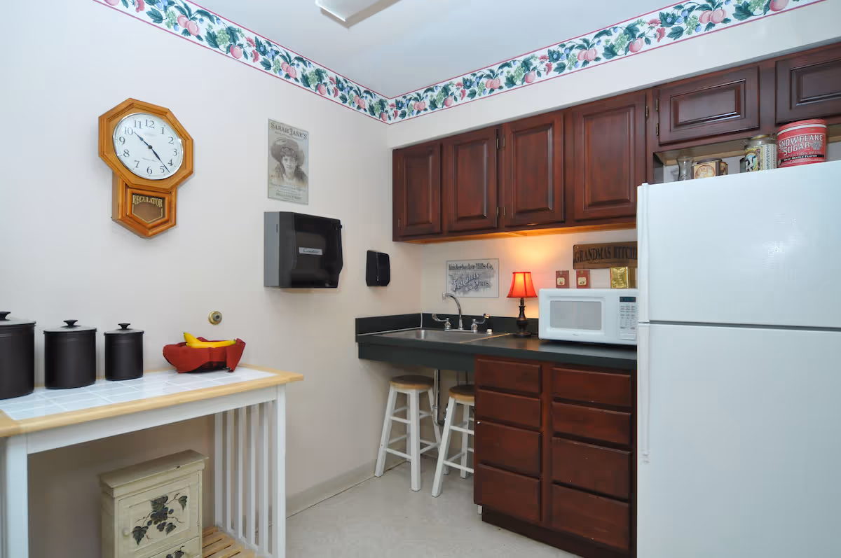 A small kitchen area with dark wooden cabinets, a white refrigerator, a microwave, a sink with two stools underneath, and a small table with three black canisters and a red basket holding bananas. The walls are decorated with a floral border near the ceiling, a wooden wall clock, and vintage-style signs. A small red lamp is placed on the countertop near the microwave.