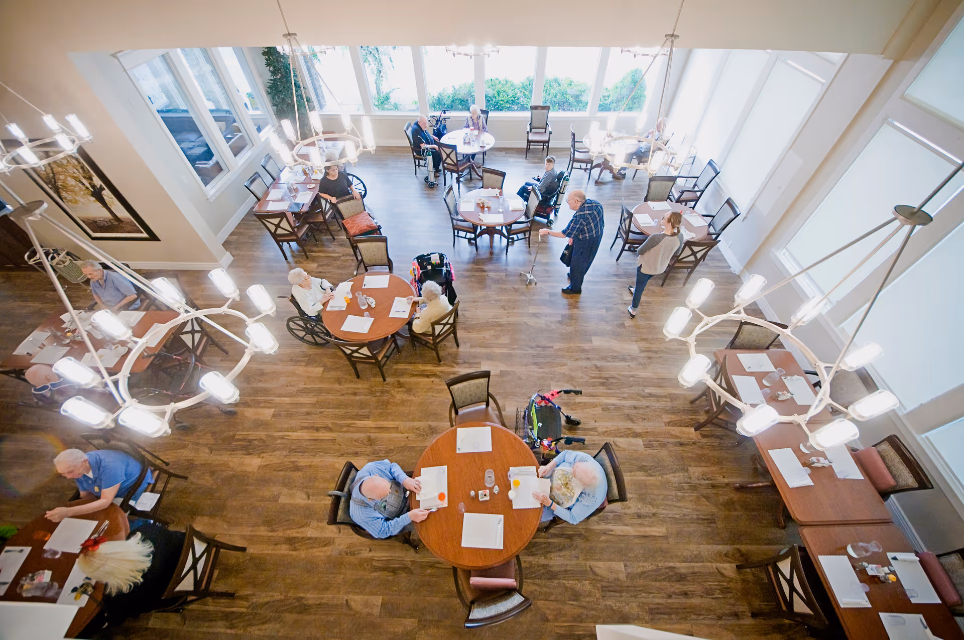 Overhead view of a dining room in a senior living facility with elderly residents seated at round and rectangular tables. Some residents are in wheelchairs, and a staff member is assisting one of them. The room has large windows, wooden floors, and modern light fixtures.