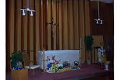 Small chapel interior with an altar decorated with flowers, a crucifix on a wood-paneled wall, chairs, and potted plants.