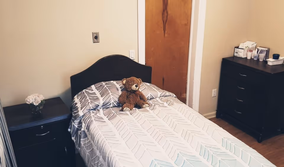 A small bedroom with a single bed covered in a patterned bedspread and a teddy bear sitting on the pillow. There is a dark wooden nightstand with a small vase of flowers on the left side of the bed and a matching dresser with various items on top on the right side. The walls are light-colored and there is a wooden door in the background.