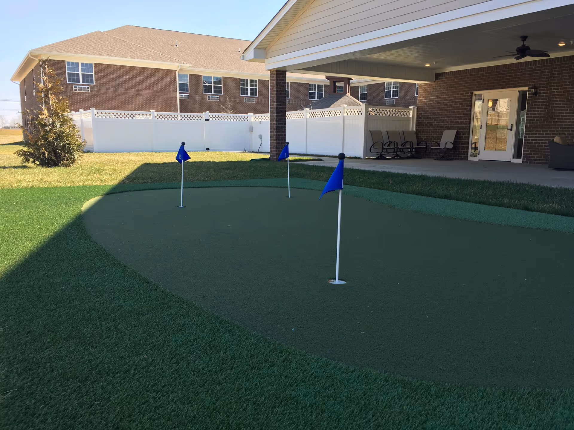 Outdoor putting green with three blue flags on holes, adjacent to a covered patio area with chairs and a ceiling fan, surrounded by a white fence and a brick building in the background.