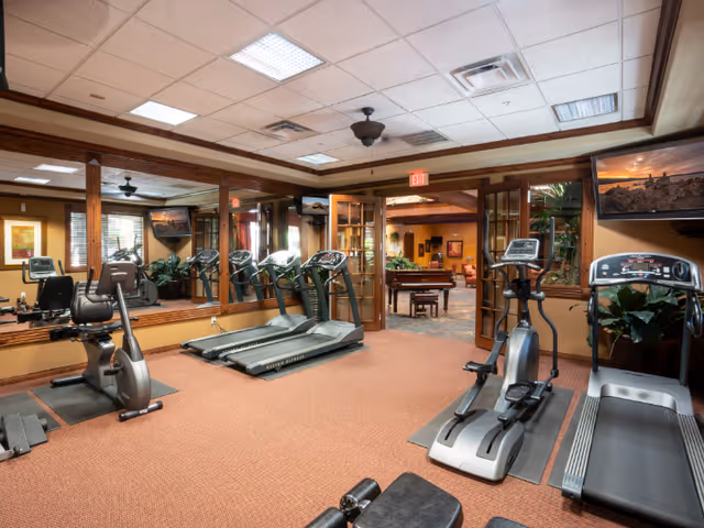 Interior view of a senior living facility fitness room with exercise equipment including treadmills, stationary bikes, and elliptical machines. The room has carpeted flooring, mirrored walls, and a ceiling with recessed lighting. Through glass doors, a lounge area with a piano and seating is visible.