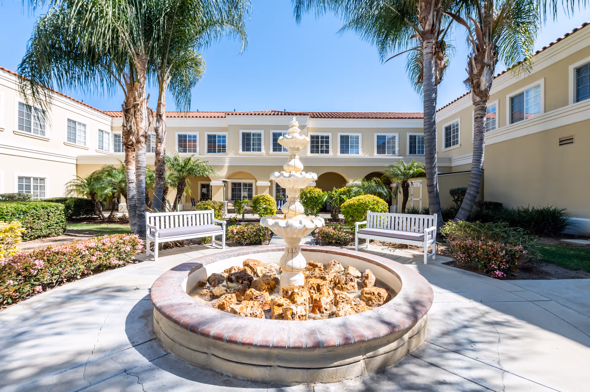 Outdoor courtyard area at San Juan Hills Healthcare Center featuring a central tiered fountain surrounded by rocks, two white benches, palm trees, bushes, and a two-story beige building with multiple windows under a clear blue sky.
