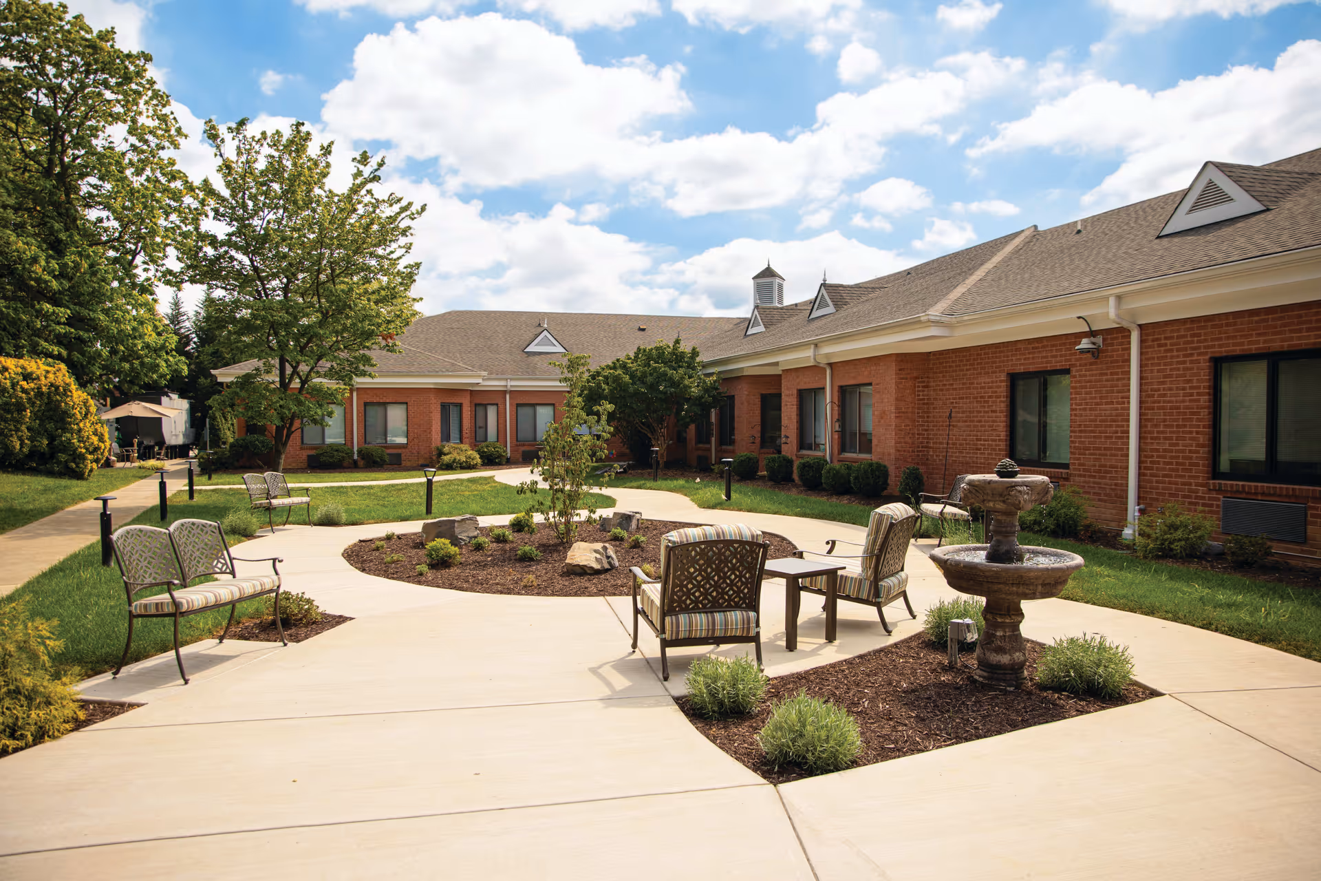 Outdoor courtyard area of a senior living facility with a circular paved walkway, garden beds with small shrubs, a two-tiered stone fountain, and several cushioned metal chairs and benches. The courtyard is surrounded by a single-story brick building under a partly cloudy sky.