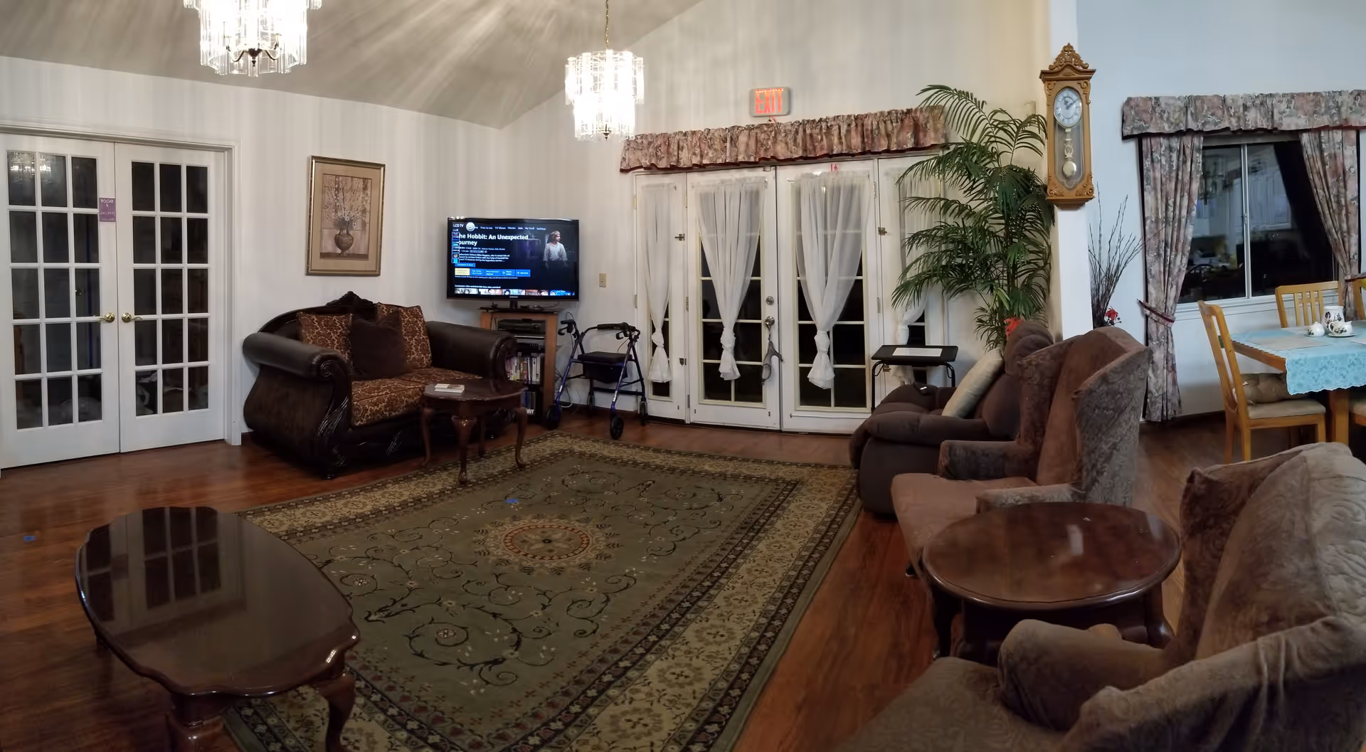 A cozy living room with multiple upholstered chairs and sofas arranged around a large patterned rug. There is a wooden coffee table in the center and a small side table. A flat-screen TV is mounted on the wall showing a movie selection screen. French doors with sheer curtains lead outside, and there is a tall grandfather clock and a large potted plant near the doors. A dining table with chairs and a lace tablecloth is visible in the background near a window with floral curtains. The room has wooden flooring and is lit by two hanging chandeliers.