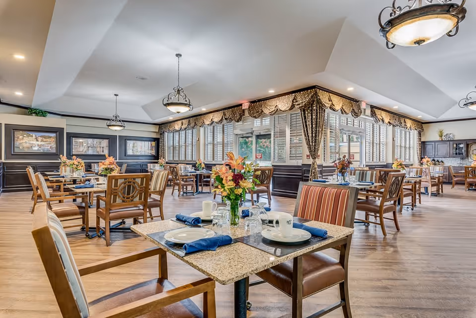 Spacious, well-lit dining room with multiple set tables, floral centerpieces, and wooden chairs.