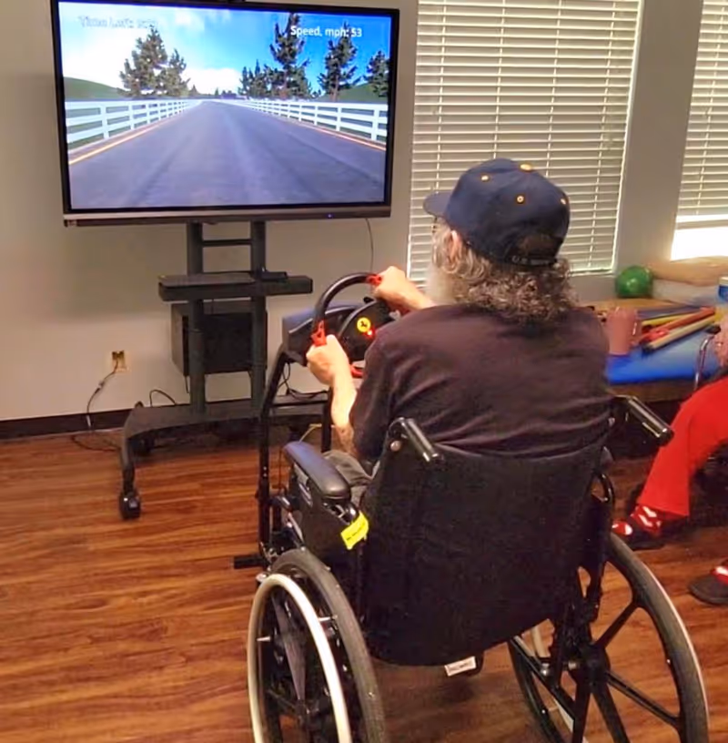 An elderly person in a wheelchair playing a driving simulation game on a large TV screen, holding a steering wheel controller in a room with wooden flooring and window blinds.