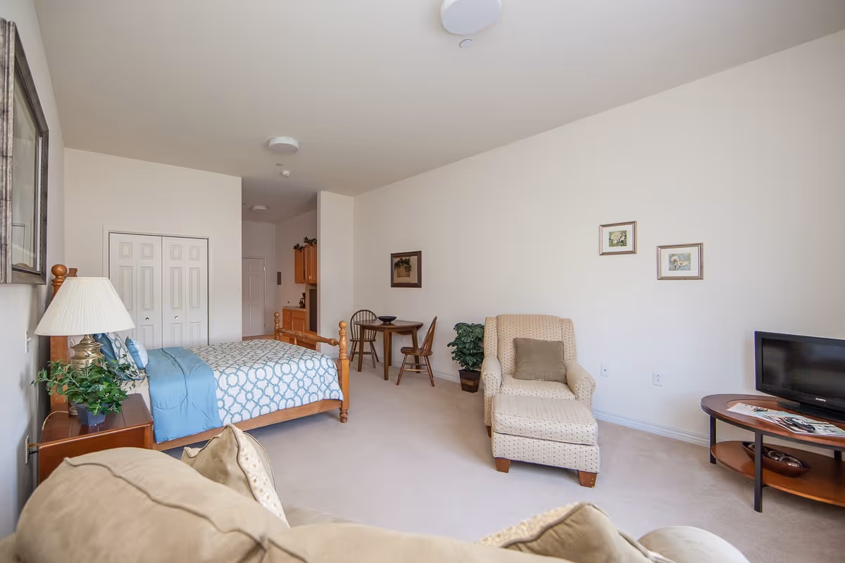 A bright and spacious senior living bedroom featuring a wooden bed with blue and white patterned bedding, a bedside table with a lamp and plant, a beige armchair with a matching ottoman, a small round table with two chairs, a flat-screen TV on a wooden stand, and framed artwork on the walls.