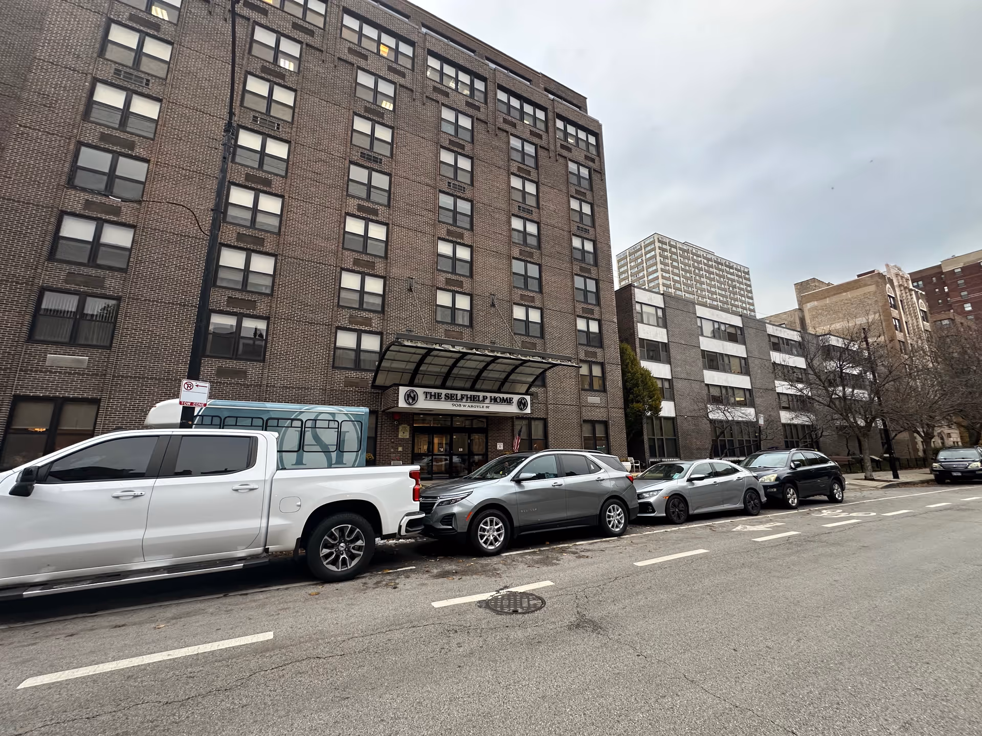 Brick multi-story senior living building with a canopy entrance labeled "The Selfhelp Home" and cars parked along the street.