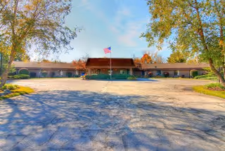 Wide exterior view of a single-story healthcare center building with an American flag on a flagpole in front, surrounded by trees and a large paved driveway under a partly cloudy sky.