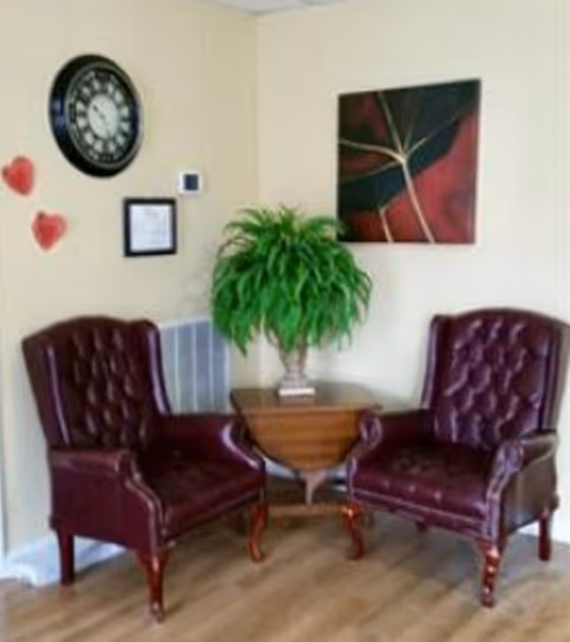 Two dark red leather armchairs with tufted backs are positioned on either side of a small wooden table holding a green potted plant. The room has light-colored walls with a large clock, two red heart decorations, a framed document, and a modern abstract painting hanging on the walls. The floor is wooden.