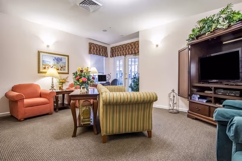 A cozy living room area in TerraBella Hillsborough featuring a striped sofa, a red armchair, a wooden coffee table with a flower arrangement, a wooden entertainment center with a TV, and a small desk with a computer near a window with valances.