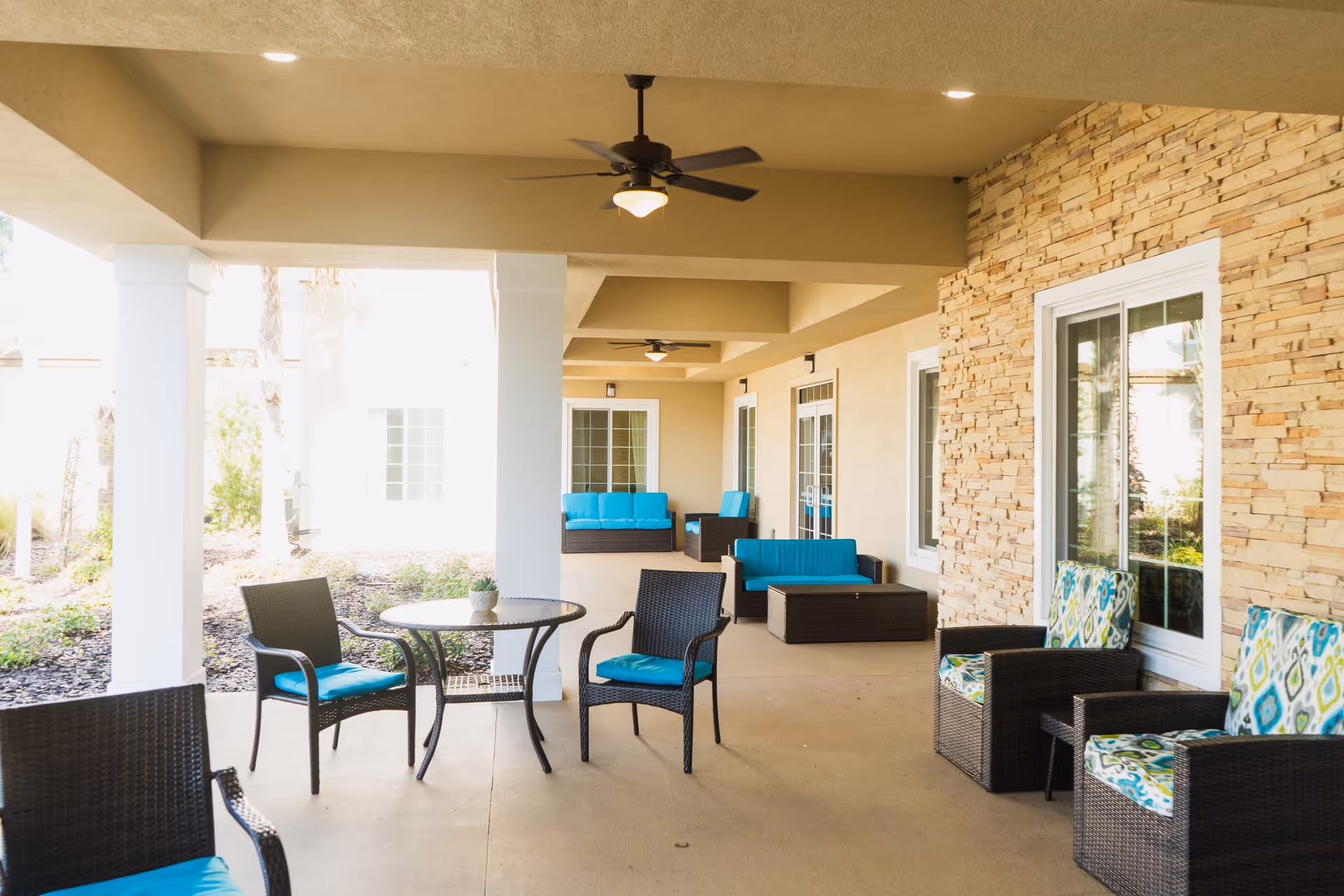 Covered outdoor patio area with wicker chairs and sofas featuring blue and patterned cushions, round glass-top table with a small plant, ceiling fans, and stone accent wall with windows and doors leading inside.
