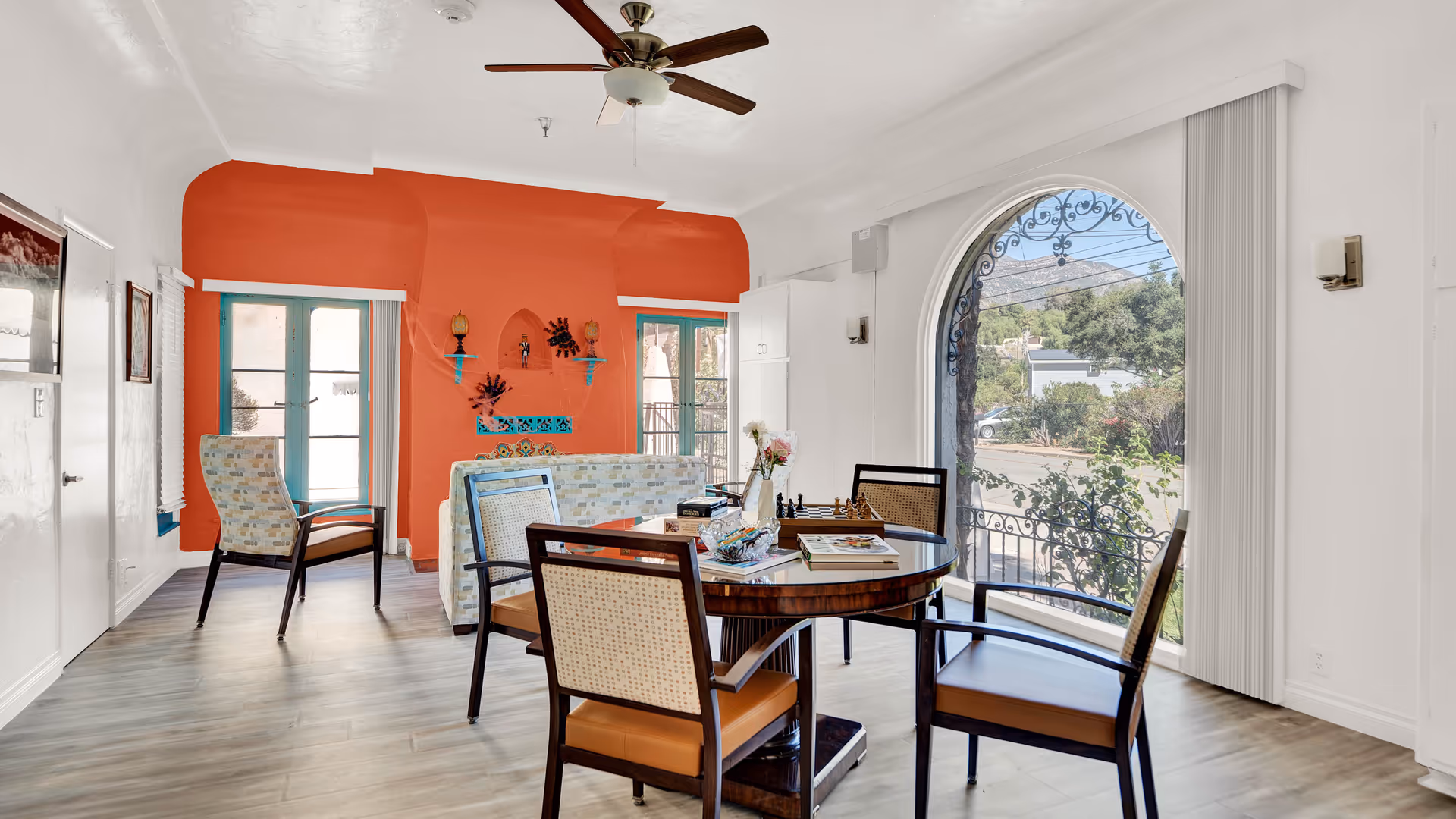 A bright and airy living room area with a round wooden table surrounded by four chairs. A chessboard, books, and a vase with flowers are on the table. Behind the table is a patterned sofa facing a wall painted in a warm terracotta color with decorative shelves and ornaments. Large arched window with vertical blinds offers a view of trees and mountains outside. The room has light wood flooring and a ceiling fan with wooden blades.