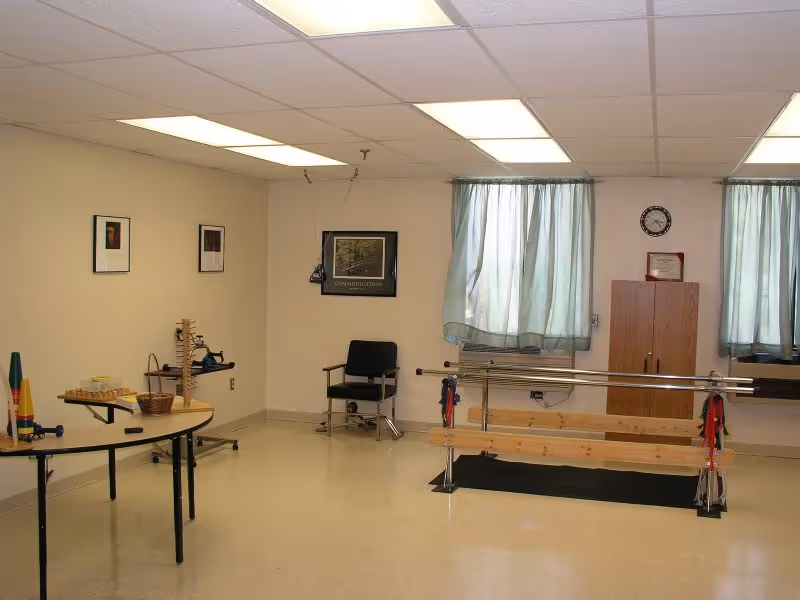 A therapy or activity room with a parallel bar setup for walking exercises, a black chair, a wooden cabinet, two windows with light green curtains, and a table with various therapy or activity items including colorful cones and wooden blocks.