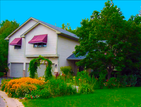 Two-story beige building with purple awnings, surrounded by a lush garden and trees.