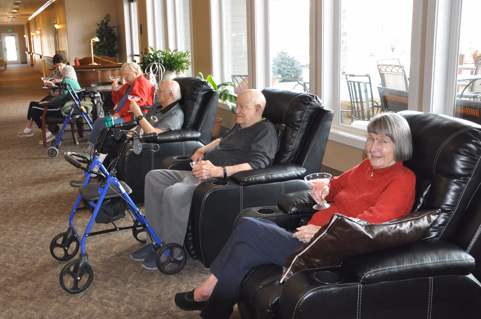 Several elderly individuals sitting in black leather recliners along a windowed wall in a well-lit common area, each holding a drink. Some walkers are positioned nearby, and there is a hallway extending in the background.