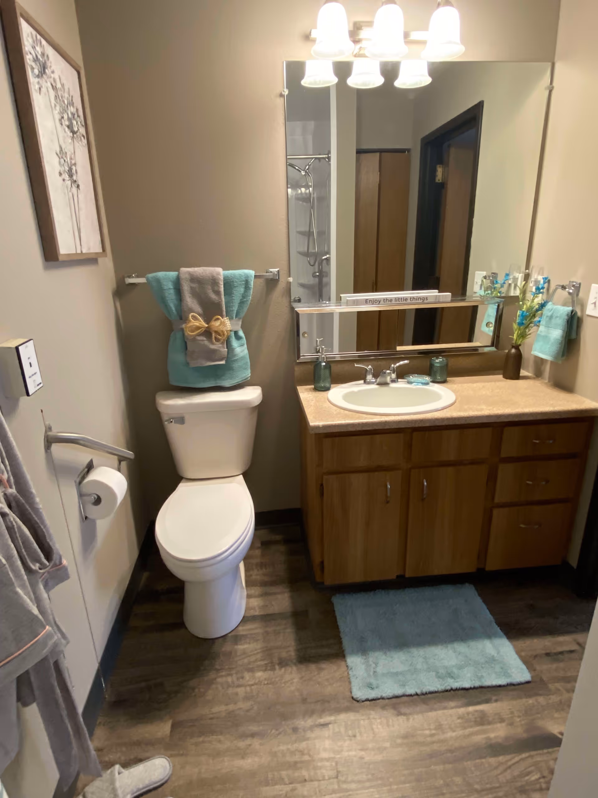 A clean bathroom with a white toilet, a wooden vanity with a sink, and a large mirror above it. The toilet has folded teal and gray towels with a decorative bow on the towel rack above it. There is a teal bath mat on the wooden floor in front of the vanity. A small vase with flowers and soap dispensers are on the vanity countertop. A framed picture hangs on the wall to the left.