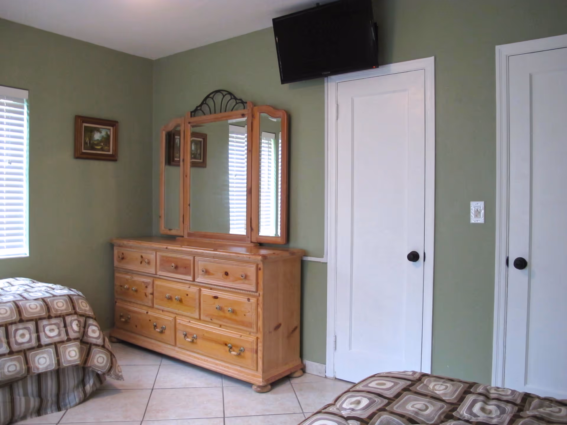 A bedroom with green walls, two beds with patterned bedspreads, a wooden dresser with a three-panel mirror, a wall-mounted TV above a white door, and a small framed painting on the wall near a window with white blinds.