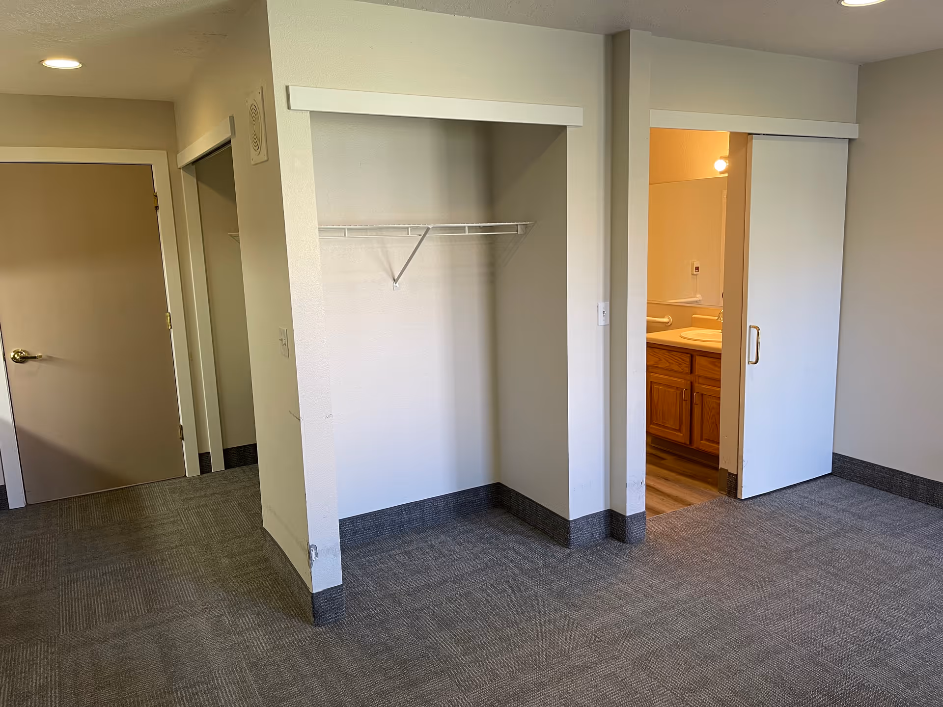 Empty assisted-living bedroom with an open closet, sliding door to a bathroom, and carpeted floor.