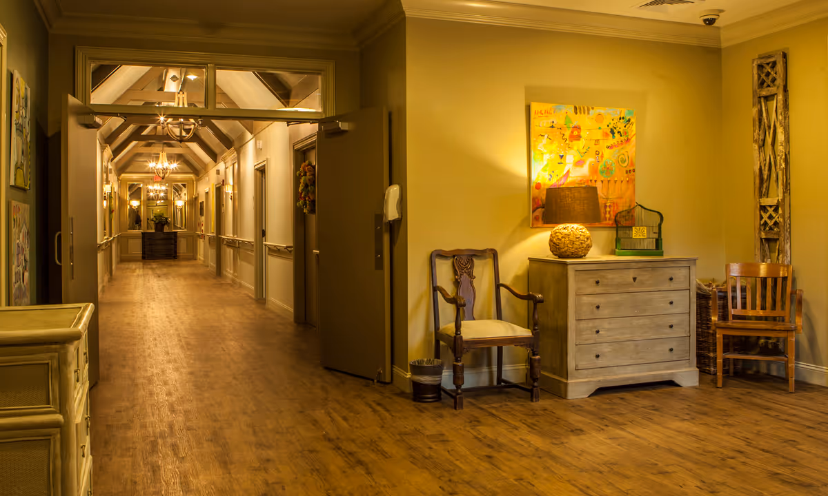 A warmly lit hallway in an assisted living community with wooden flooring, beige walls, and a vaulted ceiling with chandeliers. On the right side, there is a small sitting area with two wooden chairs, a chest of drawers with a lamp and decorative items, and artwork on the wall. The hallway extends into the distance with several doors and wall-mounted lights.