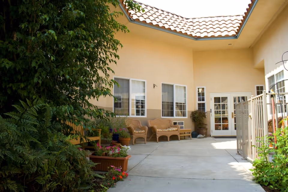 Outdoor courtyard area with beige stucco walls and a tiled roof. There are several wicker chairs with cushions arranged along the wall, potted plants and flowers placed around the space, and a gated area on the right side. The courtyard is well-lit with natural sunlight.
