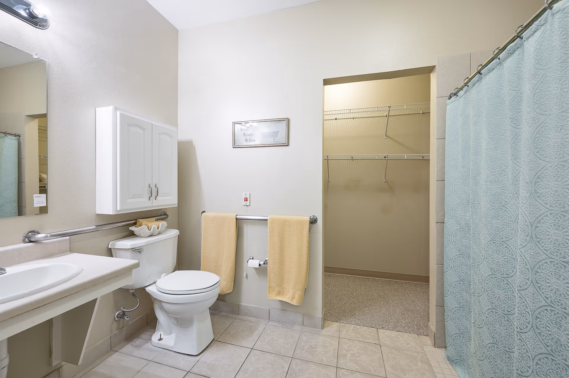 A clean bathroom with a white toilet, a sink with a countertop, a wall-mounted cabinet above the toilet, two yellow towels hanging on a towel rack, a framed picture on the wall, and a blue patterned shower curtain. There is an open doorway leading to a walk-in closet with wire shelving.