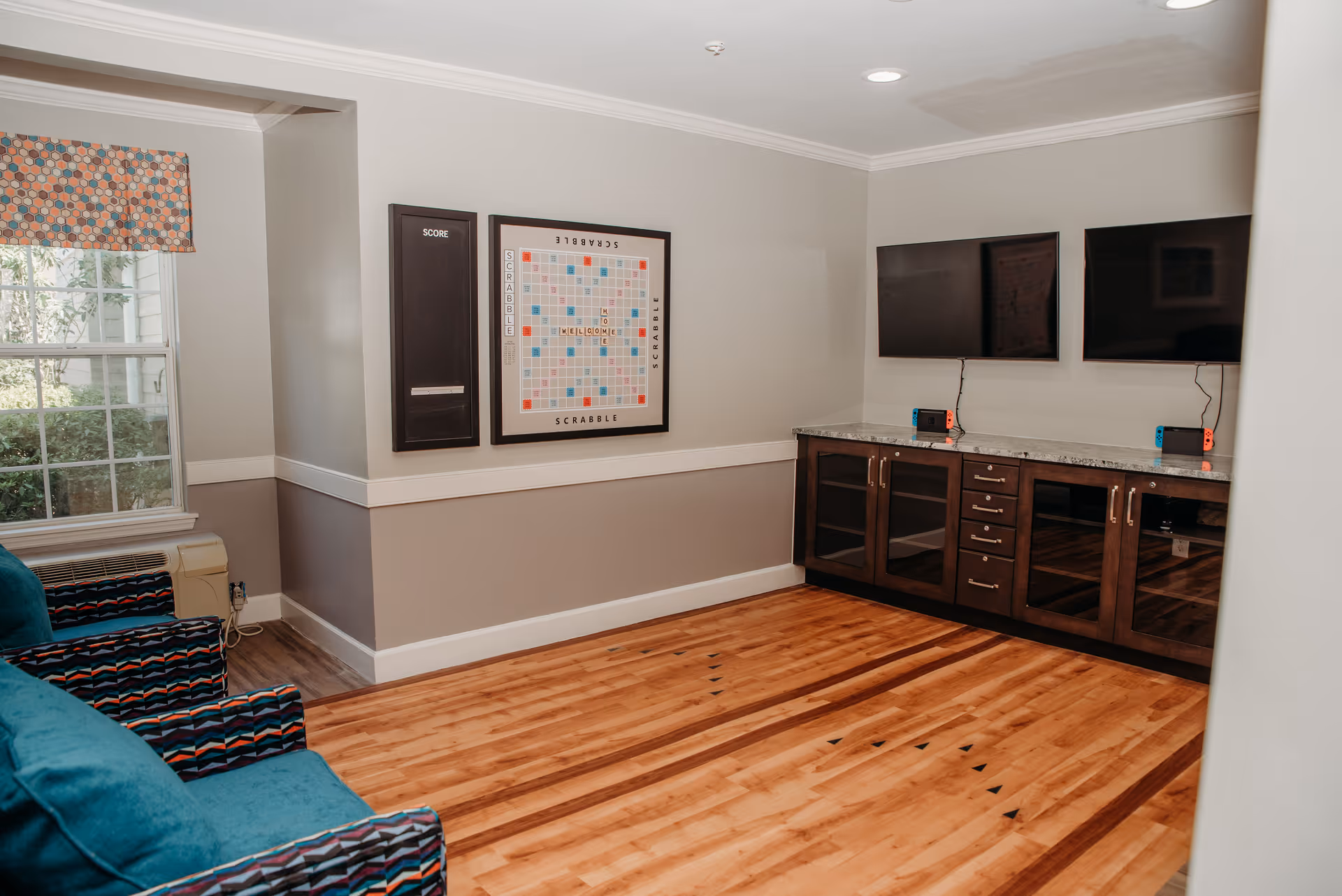 A cozy game room with wooden flooring, two teal armchairs with patterned armrests, a large window with a colorful valance, a Scrabble board mounted on the wall with a score board beside it, and a cabinet with a granite countertop holding two flat-screen TVs and two Nintendo Switch consoles.