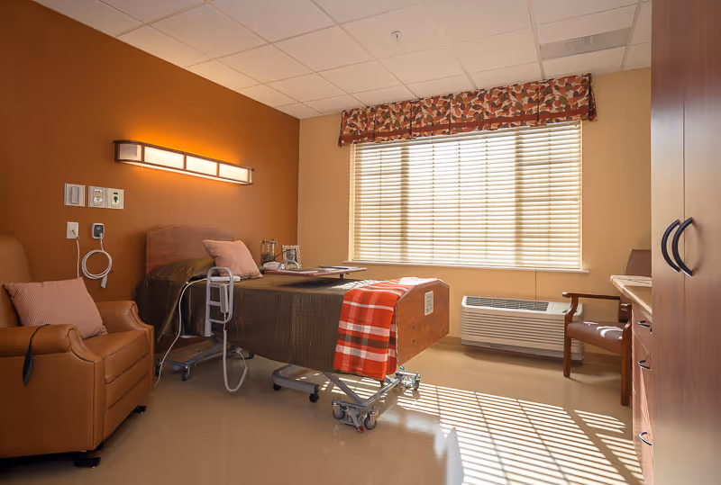 A well-lit senior care facility bedroom with a hospital-style bed covered in a brown blanket and a red plaid throw. The room has a large window with blinds and a patterned valance, a brown leather armchair with a pink pillow, a wooden chair, and a wooden cabinet. Medical equipment is attached to the wall near the bed.