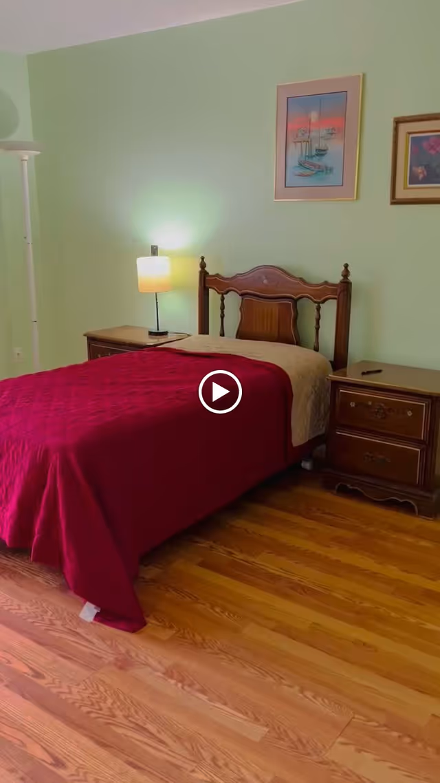 A bedroom with a wooden bed frame and a red quilted bedspread. There are two wooden nightstands on either side of the bed, one with a lamp turned on. The walls are painted light green and have two framed pictures hanging above the bed. The floor is wooden.