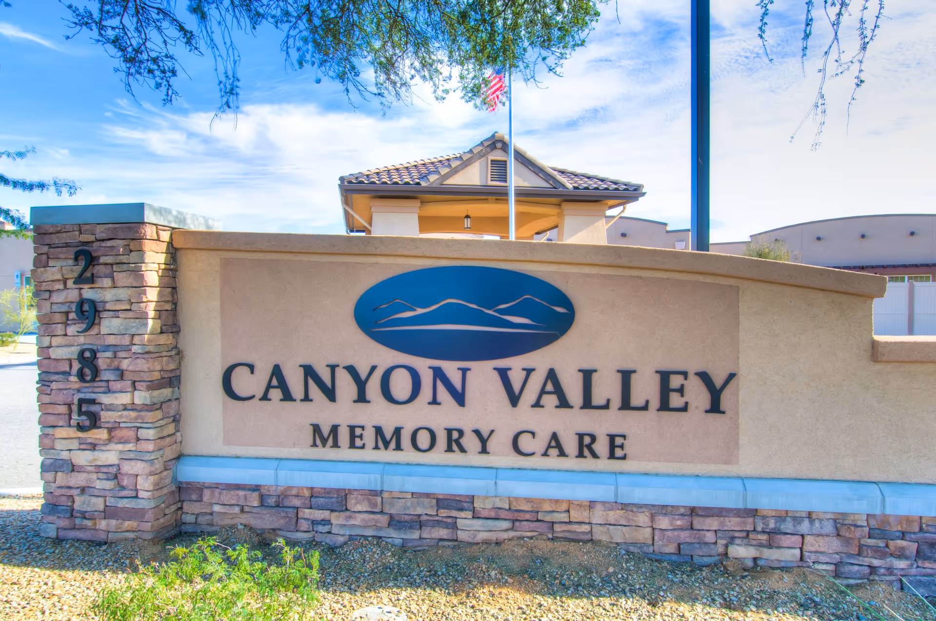 Outdoor view of a stone and stucco sign for Canyon Valley Memory Care with an address number 2985 on a stone pillar to the left. A building with a tiled roof and an American flag on a flagpole are visible in the background under a blue sky with some clouds.