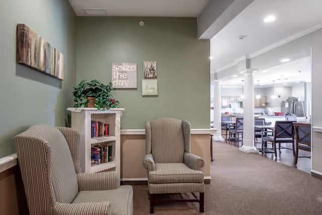 A cozy sitting area with two striped armchairs facing each other, a small white bookshelf filled with books, and a green wall adorned with three decorative wall hangings including one that says 'seize the day'. In the background, there is a spacious dining area with tables and chairs, white columns, and bright lighting.