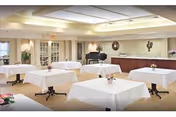 A spacious dining room with several square tables covered in white tablecloths, each adorned with a small floral centerpiece. The room features soft lighting from a large ceiling fixture, a piano against the far wall, and a counter area with decorative items and a mirror above it.