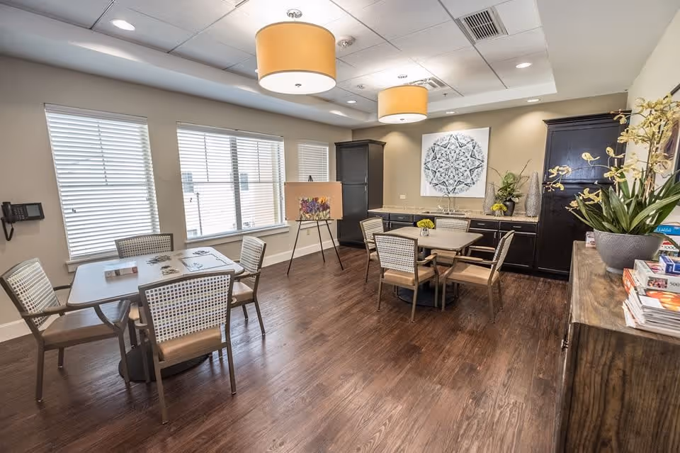 Bright communal dining/activity room with two tables and chairs, hardwood floors, large windows with blinds, cabinetry and wall art.