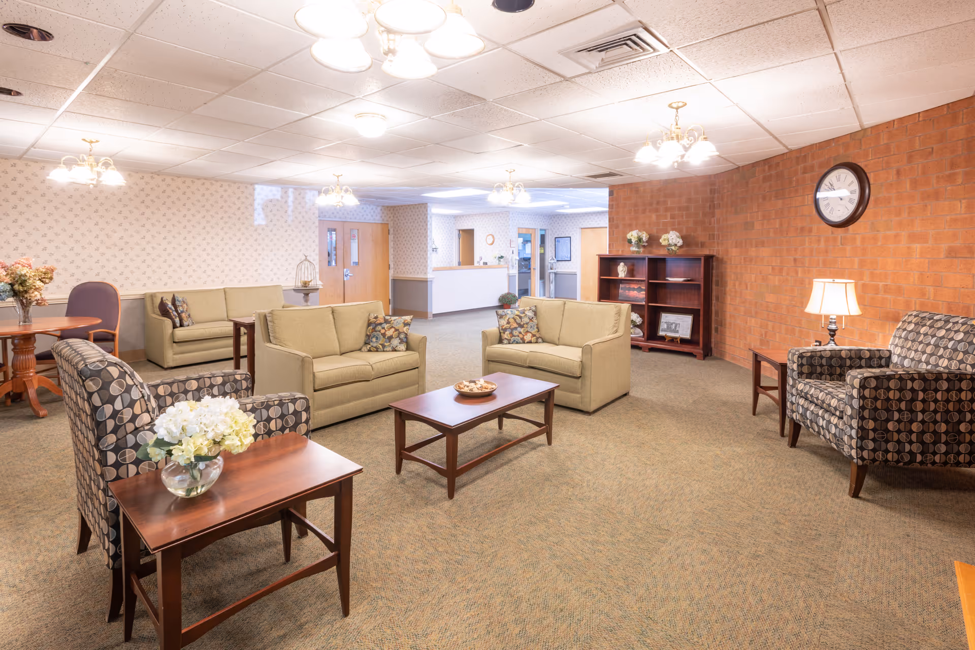 A spacious and well-lit common area in a senior living facility featuring two beige loveseats with patterned pillows, two patterned armchairs, wooden coffee and side tables with flower arrangements, a wooden bookshelf against a brick wall, a clock, and multiple ceiling light fixtures. The background shows a reception desk and doors leading to other rooms.