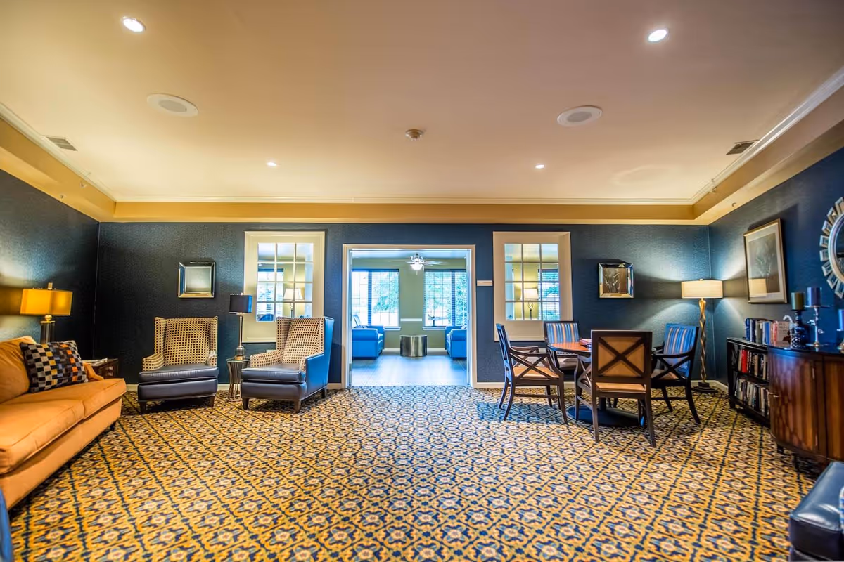 A spacious, well-lit common area with patterned carpet, featuring a tan sofa with a checkered pillow, two patterned armchairs, a round table with four chairs, and a bookshelf with decorative items and books. The walls are dark blue with framed artwork and mirrors. Through a wide doorway, another seating area with blue sofas and large windows is visible.