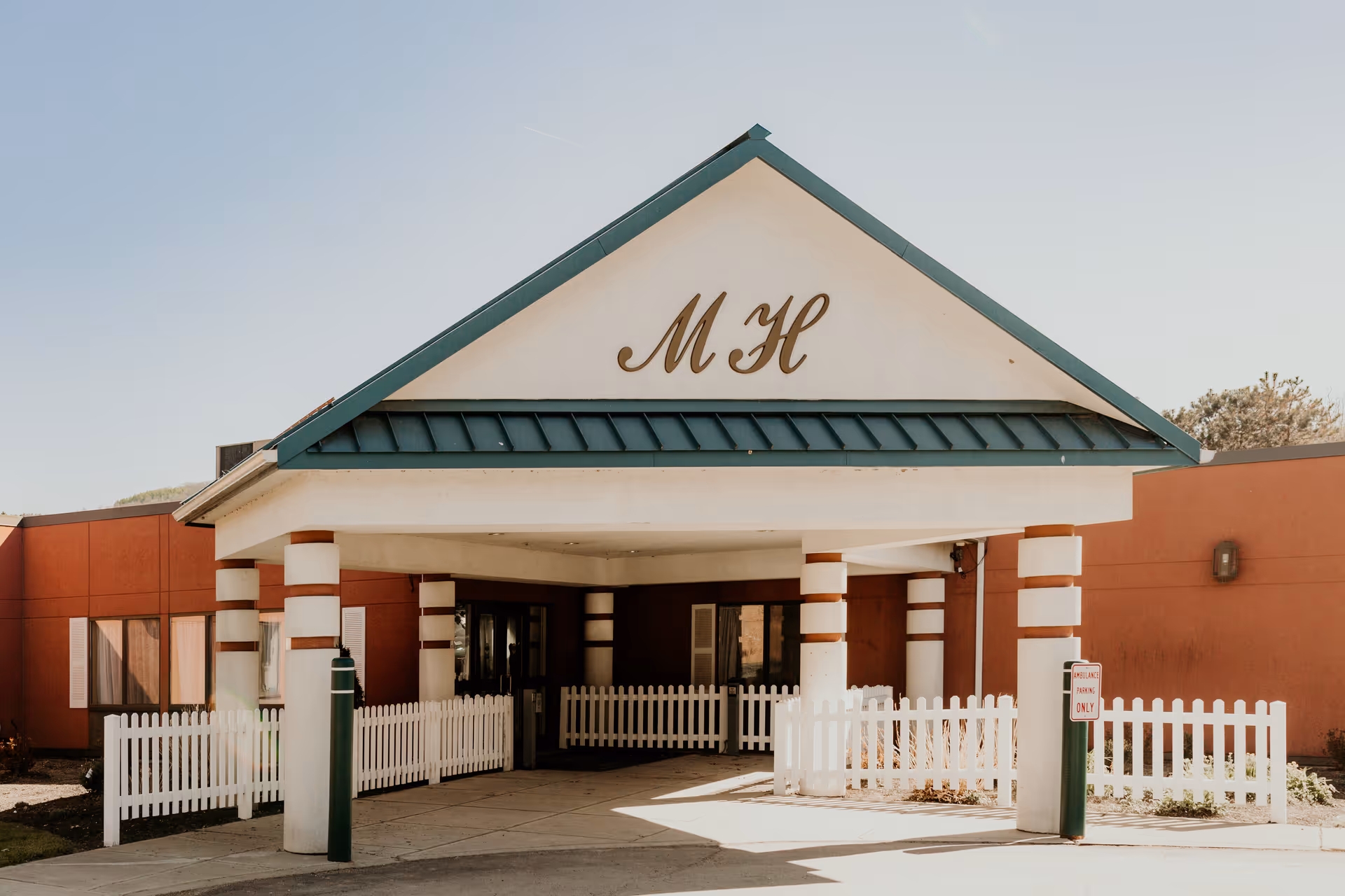 Entrance of Manor Hills: A Willow Ridge Senior Living Community with a covered driveway supported by white columns with brown stripes, white picket fences on both sides, and the letters 'MH' displayed prominently on the front gable.