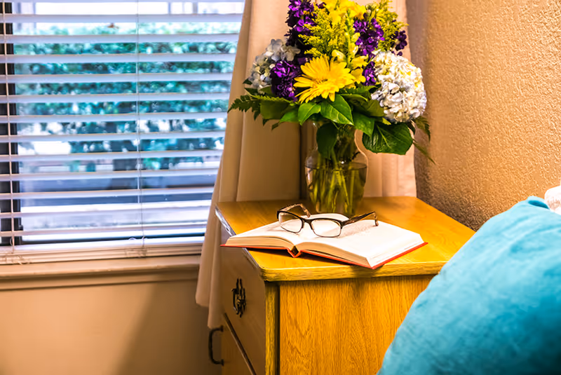 A wooden bedside table next to a window holds an open book with reading glasses and a vase of flowers.