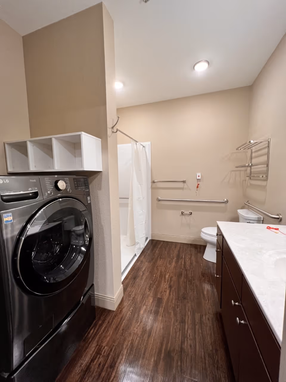 A bathroom with a dark wood floor, beige walls, and recessed ceiling lights. On the left side, there is a black front-loading washing machine with a white cubby shelf above it. In the background, there is a shower with a white curtain. On the right side, there is a toilet with grab bars on the wall, a towel rack above it, and a white countertop with dark brown cabinets underneath.