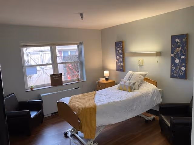 A small, tidy bedroom with a single hospital-style bed covered with a white blanket and a mustard yellow throw. There are two black armchairs on either side of the bed, a wooden nightstand with a lamp, and two floral paintings on the wall. A window with a view of an exterior building is on the left side of the room.