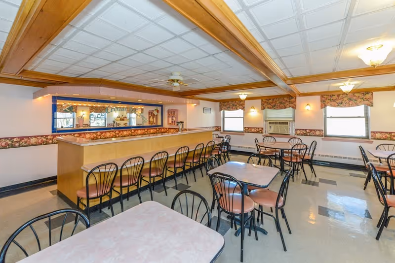 A dining room with multiple tables and chairs arranged neatly. There is a long counter with bar stools along one side, floral wallpaper border, windows with floral valances, and ceiling lights. The room has a tiled floor and a ceiling with wooden beams.
