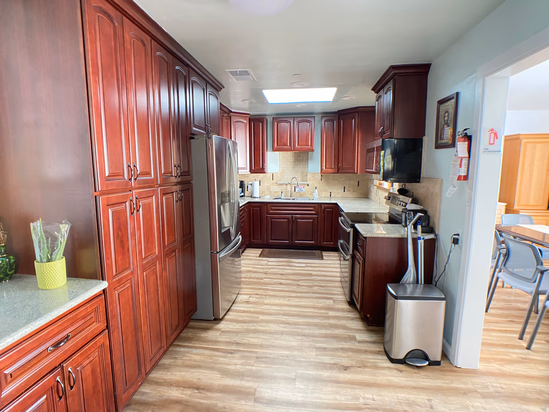 A spacious kitchen with wooden cabinets and drawers in a rich reddish-brown finish. The kitchen features a stainless steel refrigerator, stove, microwave, and a sink under a skylight. The floor is light wood, and there is a trash can and cleaning tools near the entrance. Adjacent to the kitchen is a dining area with tables and chairs visible through an open doorway.