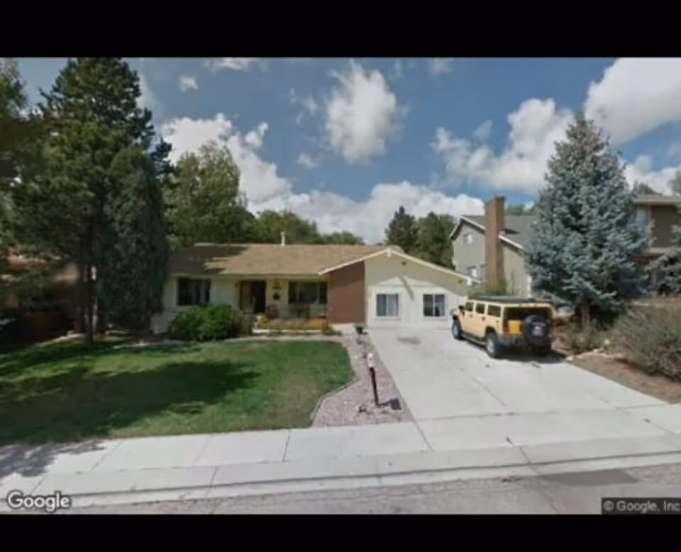 Front exterior view of a single-story house with a driveway, a yellow SUV parked on the driveway, a green lawn, trees, and a partly cloudy sky.