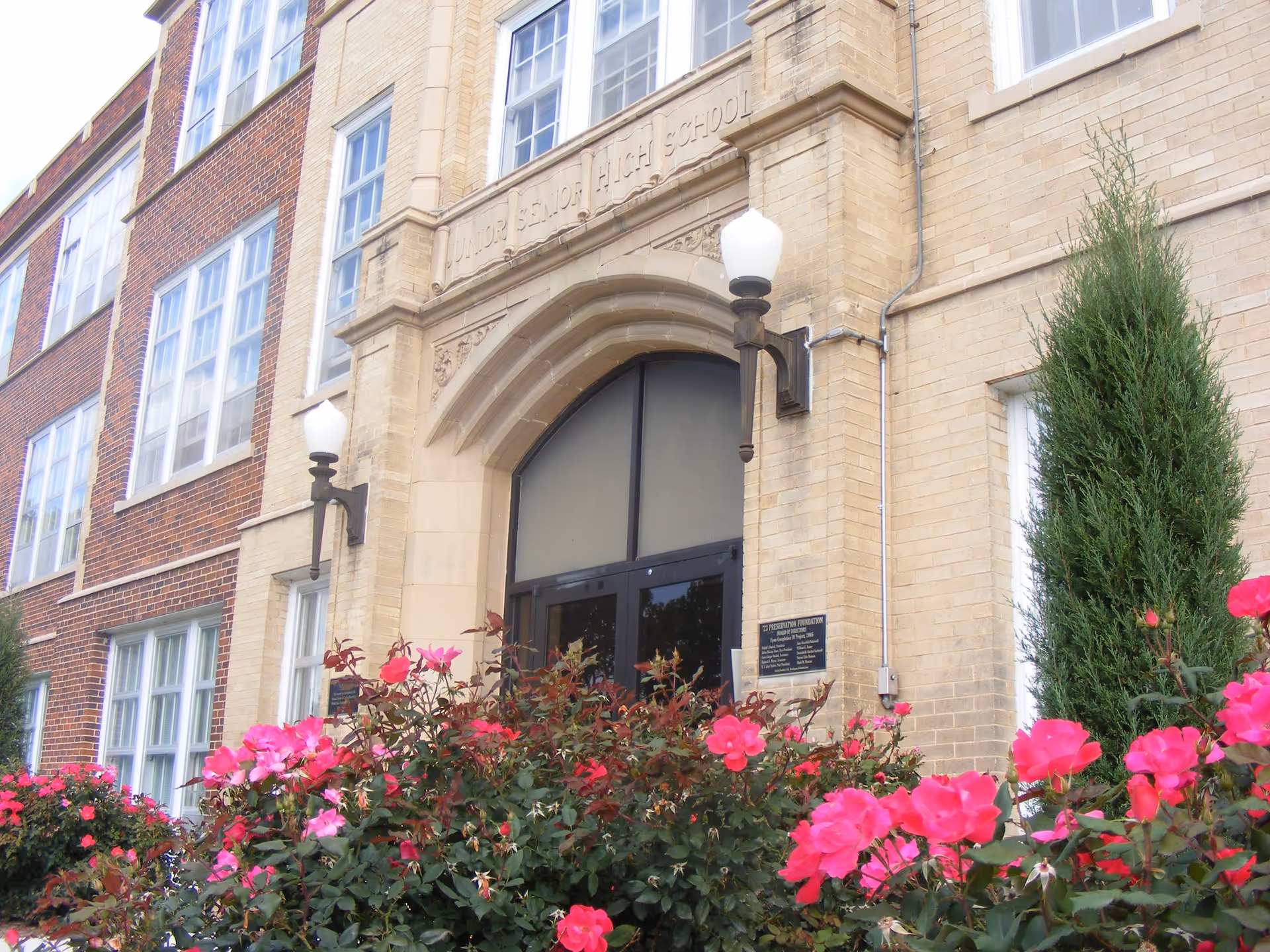 Entrance of a brick building with an arched doorway labeled 'Junior Senior High School' above it, flanked by two wall-mounted lamps. In front of the entrance, there are vibrant pink flowers and green shrubs.