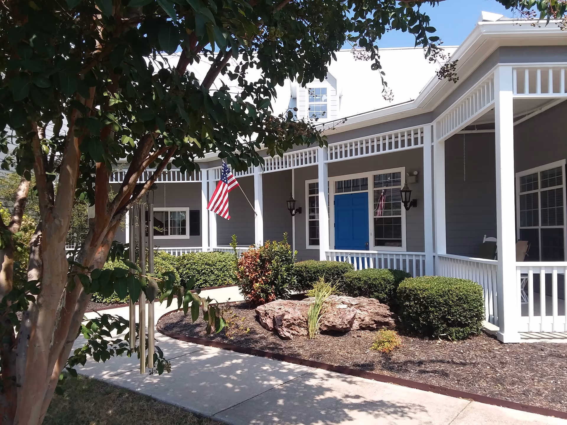Exterior view of a senior living facility with a covered porch featuring white railings and columns. There is a blue door, several windows, and an American flag mounted on the porch. The landscaping includes bushes, a tree, and a large rock in a mulched garden bed next to a concrete walkway.