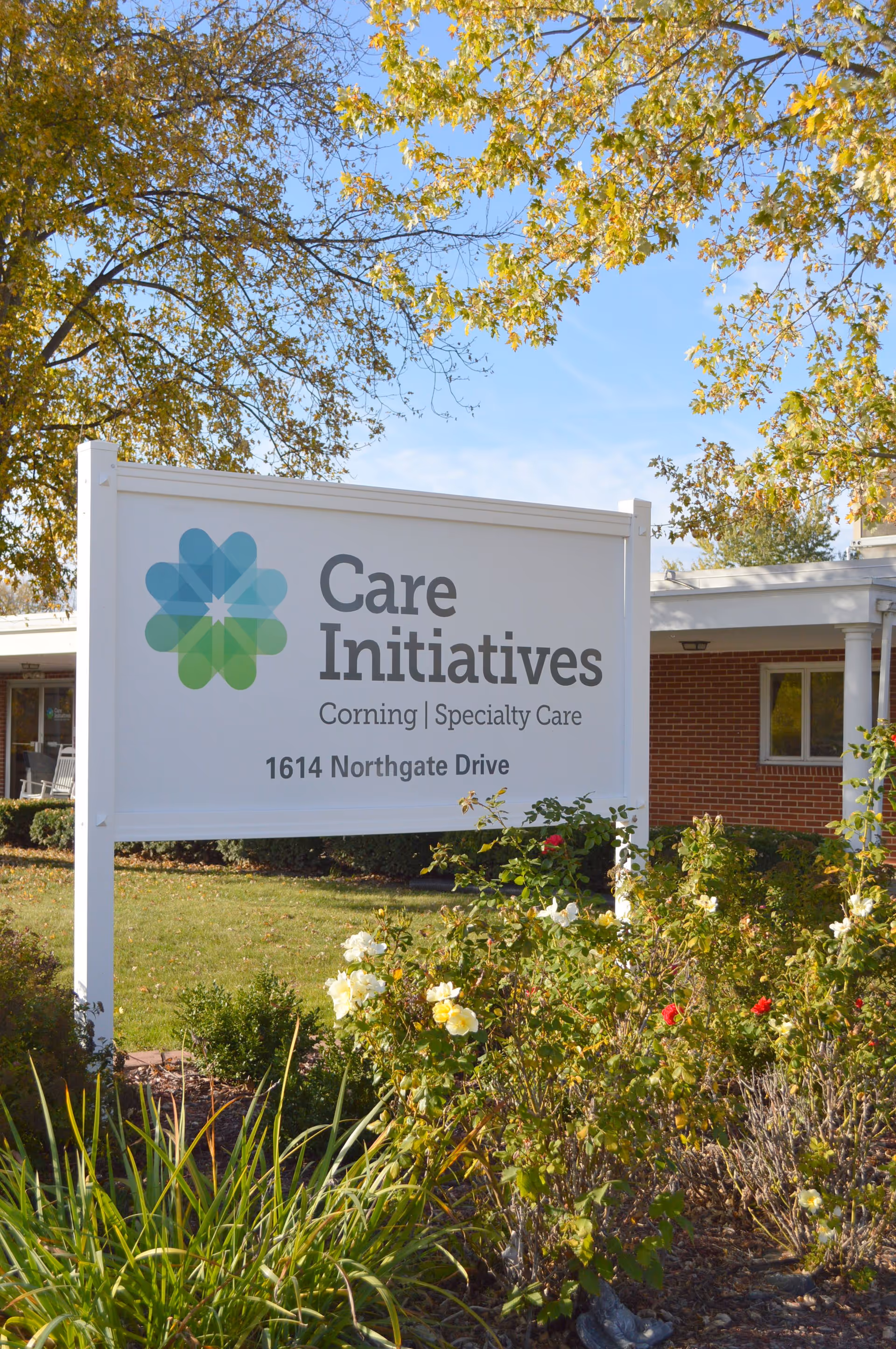 Outdoor view of a white sign for Care Initiatives Corning Specialty Care at 1614 Northgate Drive, surrounded by green bushes and flowering plants with a brick building and trees in the background under a clear blue sky.