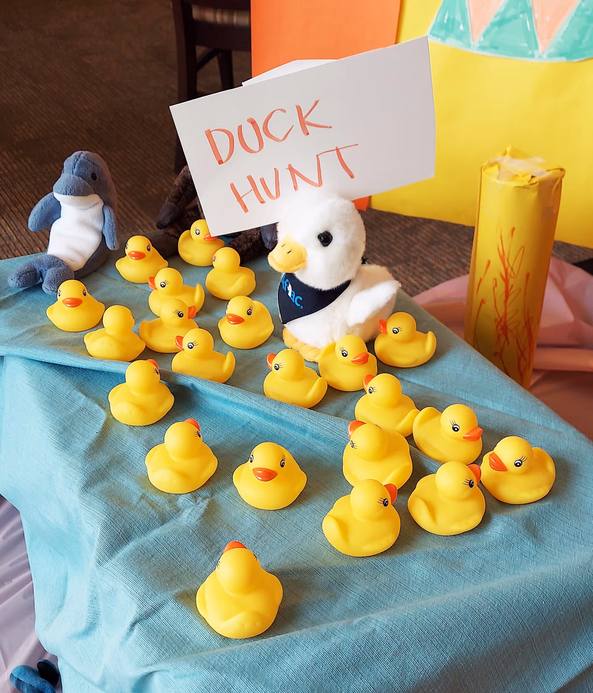 A group of yellow rubber ducks arranged on a blue cloth with a white plush duck wearing a blue bandana holding a sign that says 'DUCK HUNT'. There is also a plush dolphin and a yellow cylindrical object with red scribbles in the background.