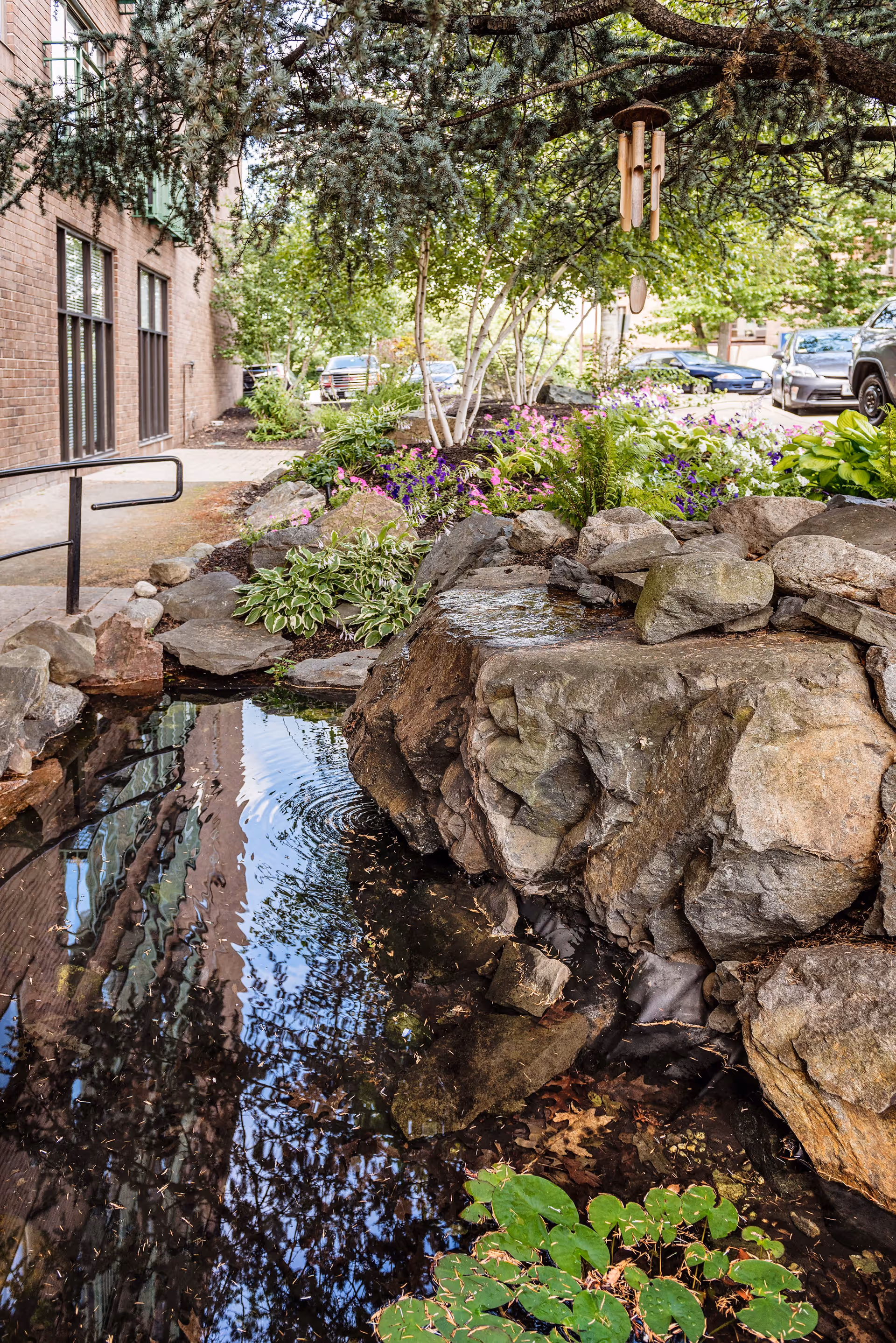 A serene outdoor garden area at AVIVA Country Club Heights featuring a small pond with rocks surrounding it, lush green plants, colorful flowers, and a wind chime hanging from a tree branch. A brick building and parked cars are visible in the background.